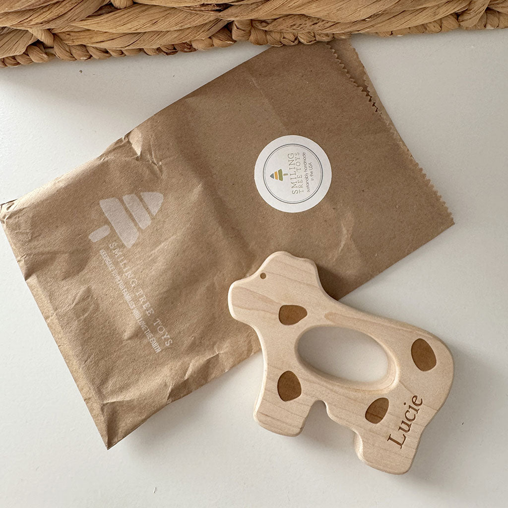 Wooden cow shaped teething toy with 'Lucie' engraving on a brown paper bag.